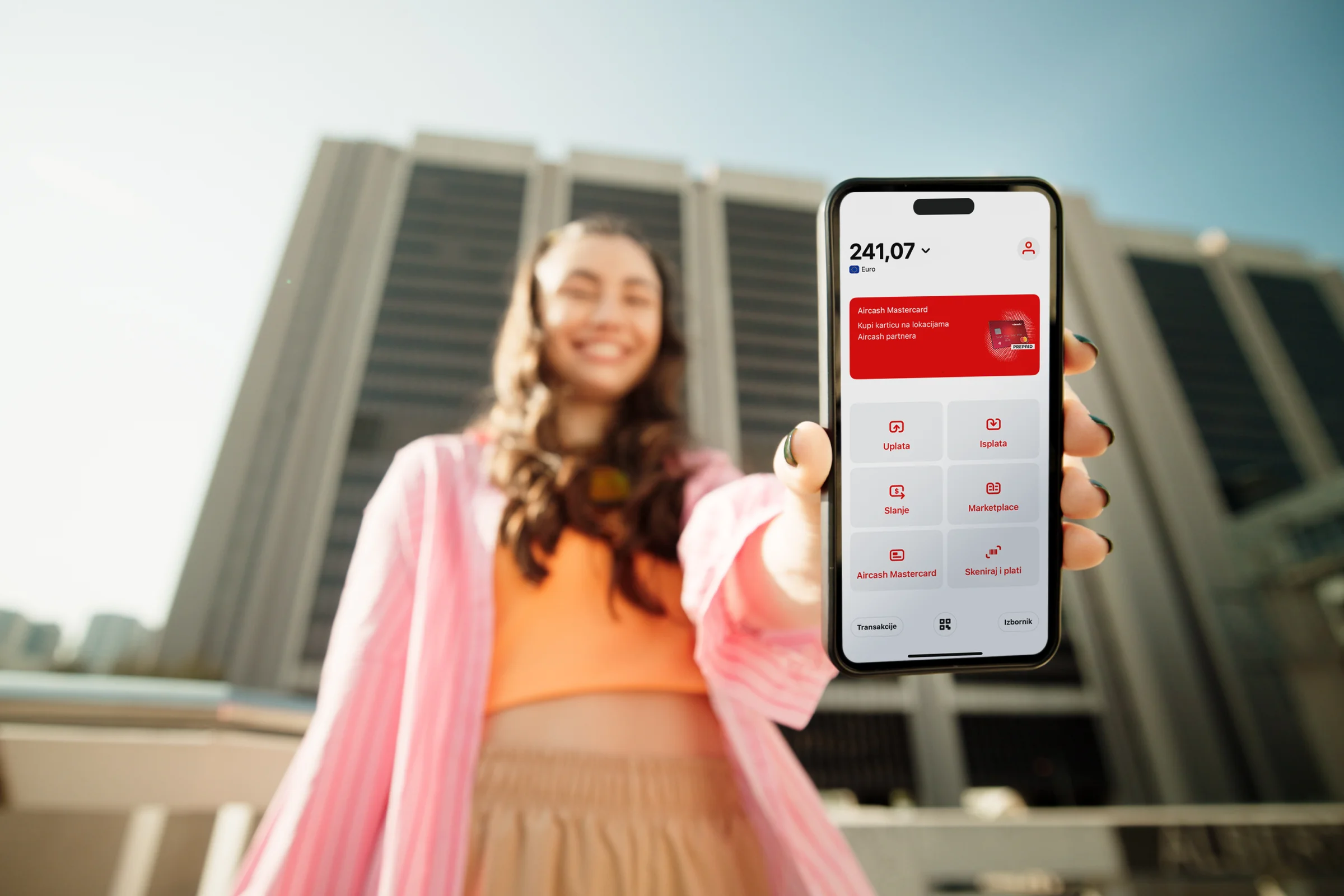 Smiling woman outdoors holding a smartphone displaying a financial app with a balance of 241.07.