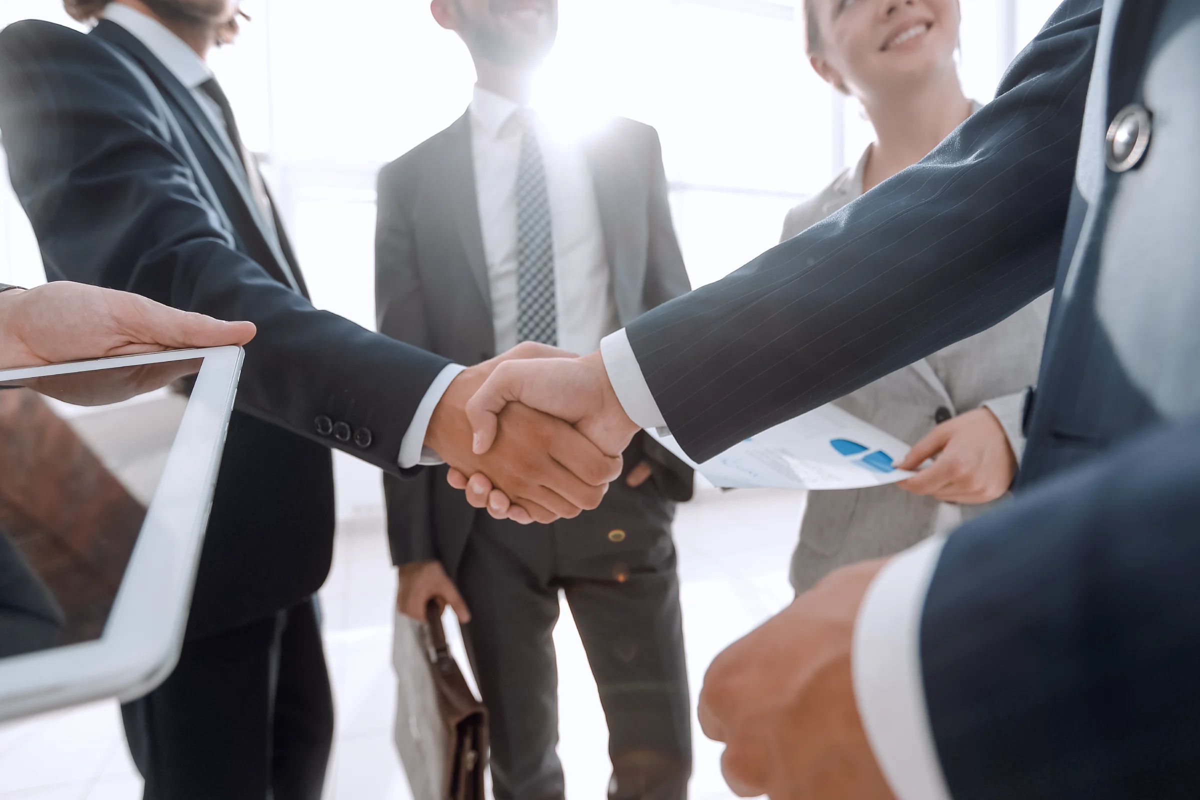 Business professionals in formal attire shaking hands, with others holding documents and a tablet, standing in a bright office setting.