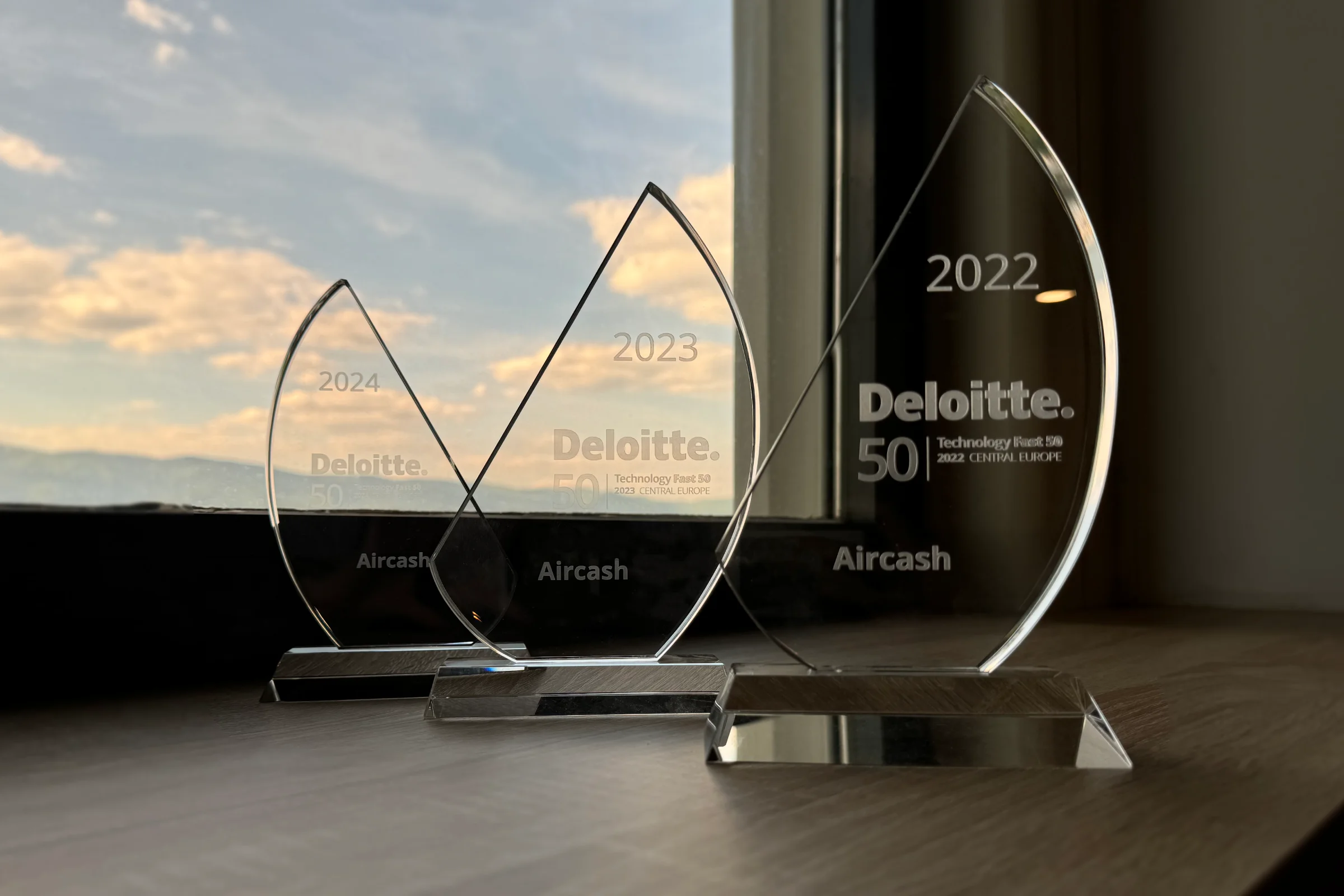 Three transparent awards with "Deloitte 50" and "Aircash" engravings for 2022, 2023, and another year, placed on a wooden surface by a window.