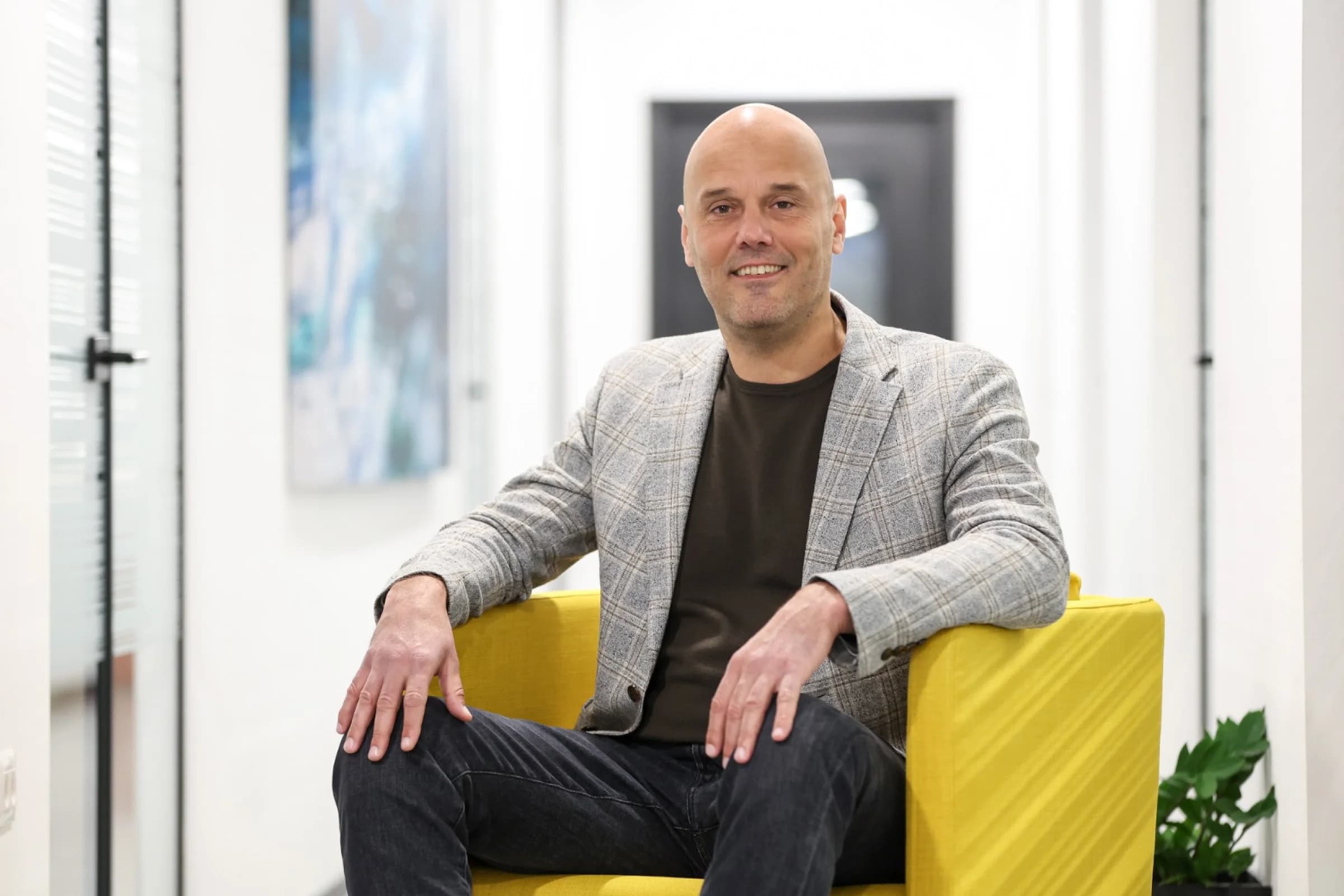 Man sitting on a yellow chair in a hallway, wearing a gray plaid blazer and dark pants, smiling at the camera.
