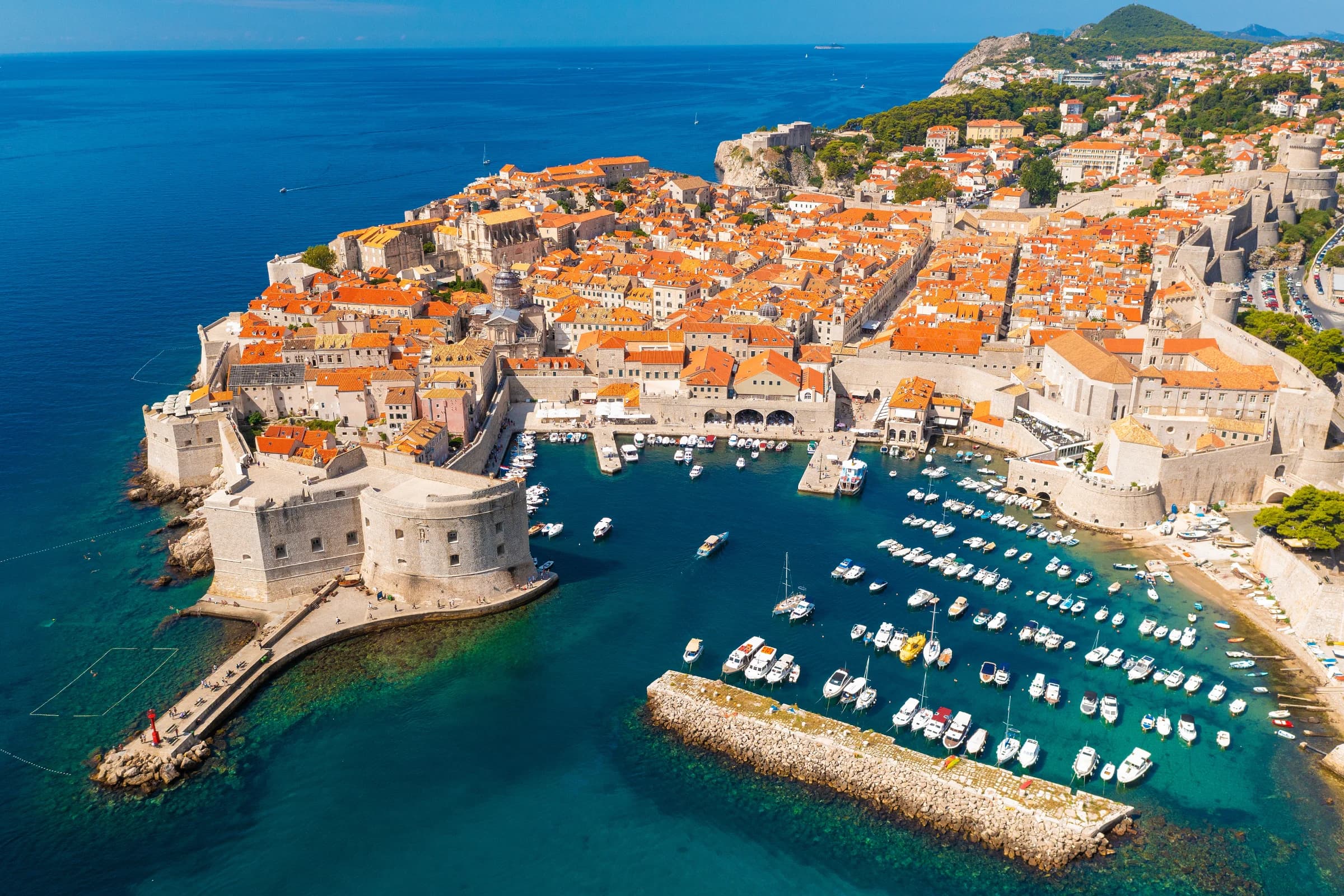 Aerial view of Dubrovnik's old town with orange rooftops, historic walls, and a harbor filled with boats against a blue sea backdrop.
