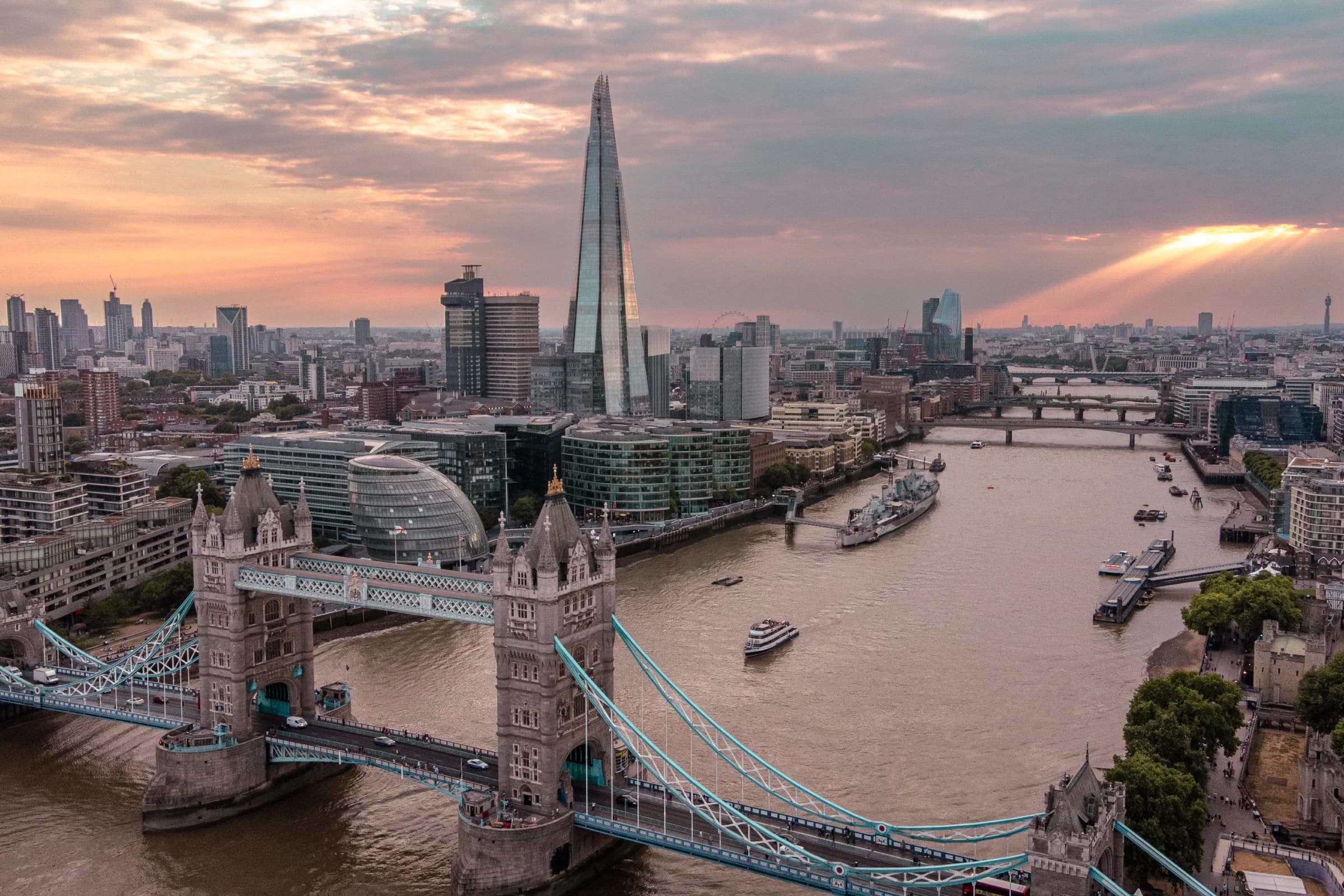 Aerial view of London featuring Tower Bridge, the Shard skyscraper, and the Thames River at sunset with a cloudy sky.