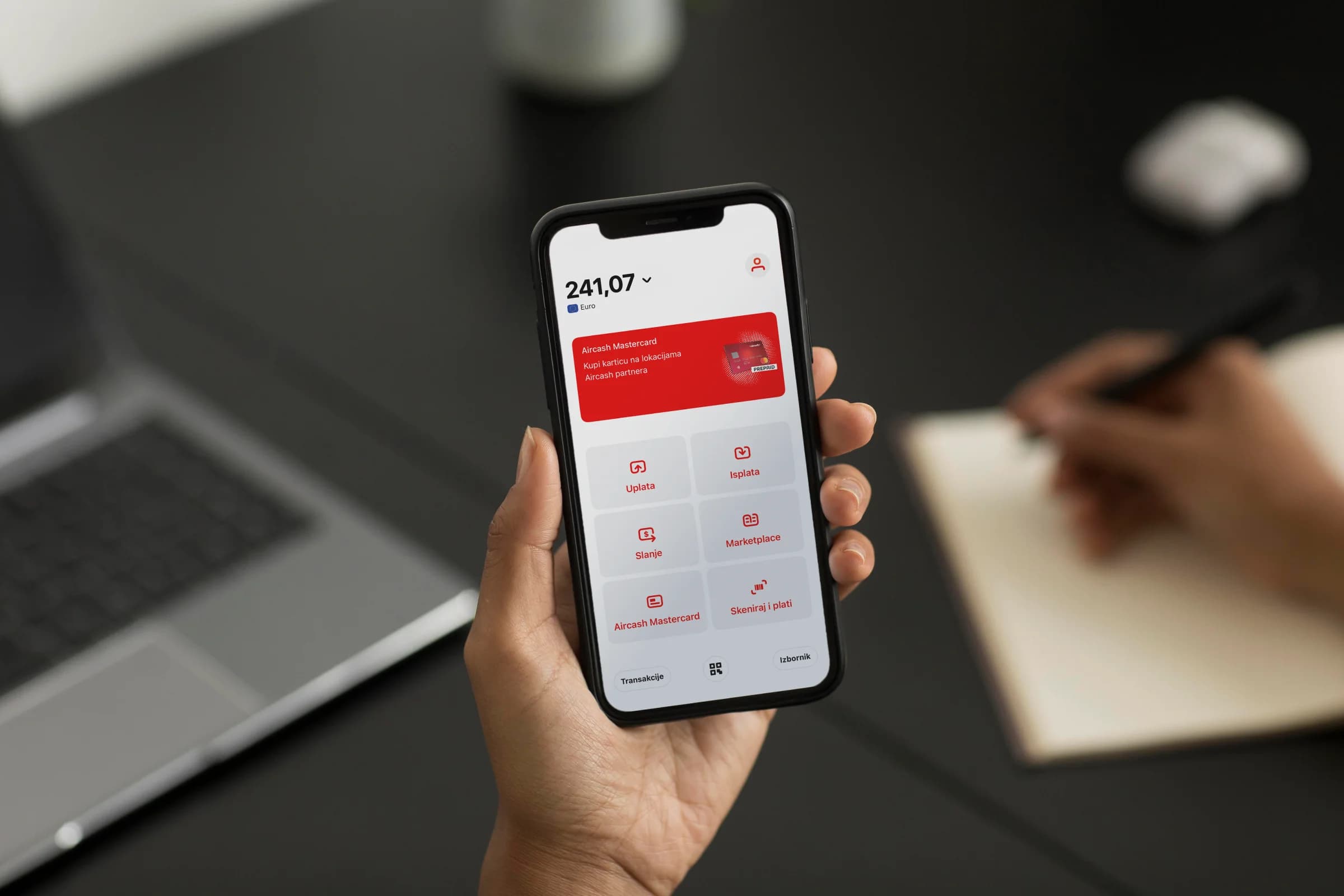 Person holding a smartphone displaying a mobile banking app with a balance of 241.07. Nearby, another hand writes in a notebook.