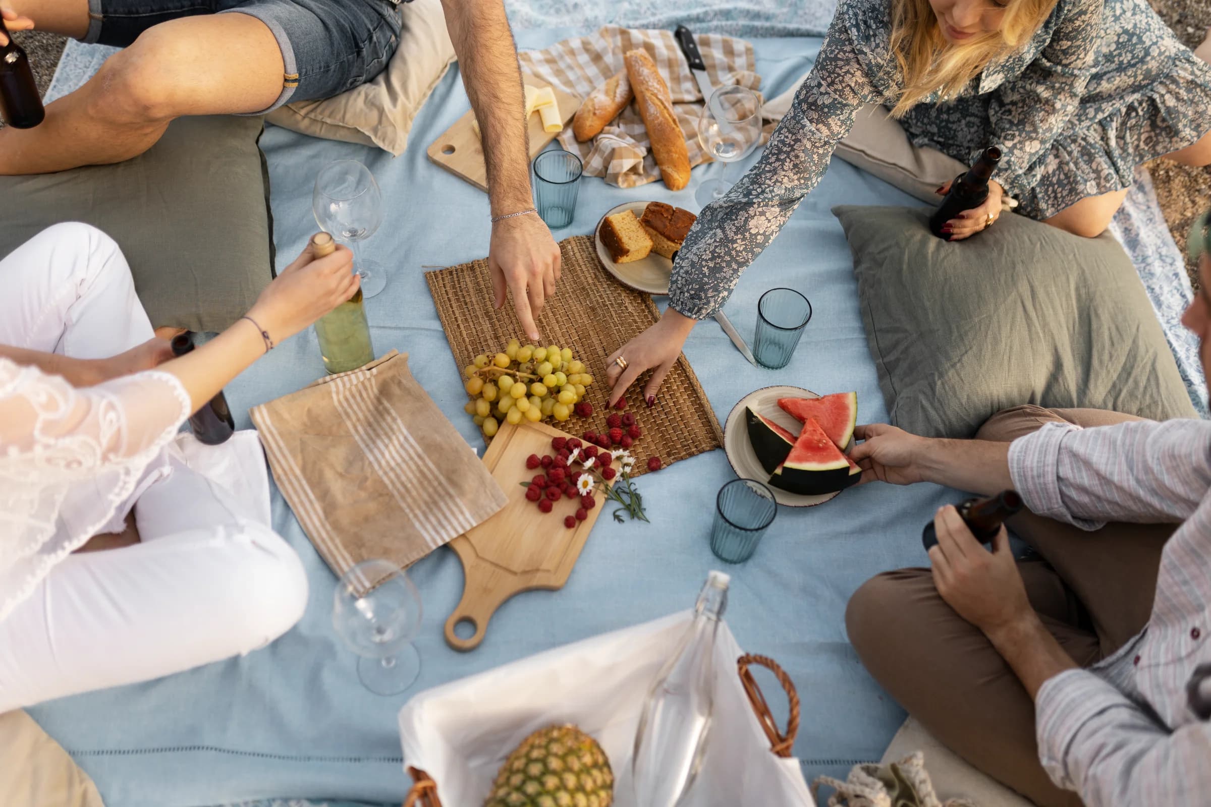 People enjoying a picnic on a blue blanket with fruits, bread, and drinks. Hands are reaching for food items.