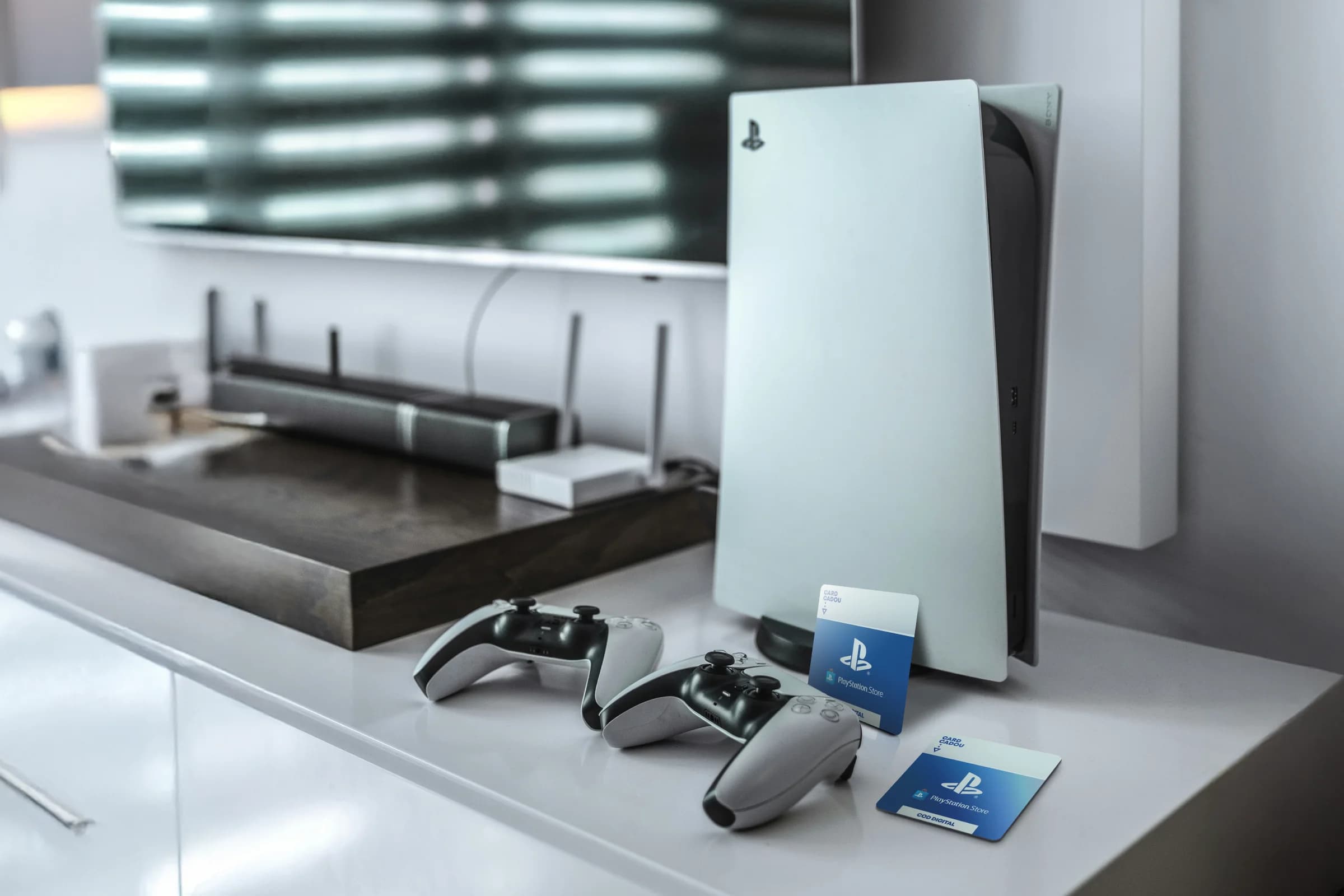 A PlayStation 5 console with two controllers on a white shelf, next to a TV and PlayStation gift cards.