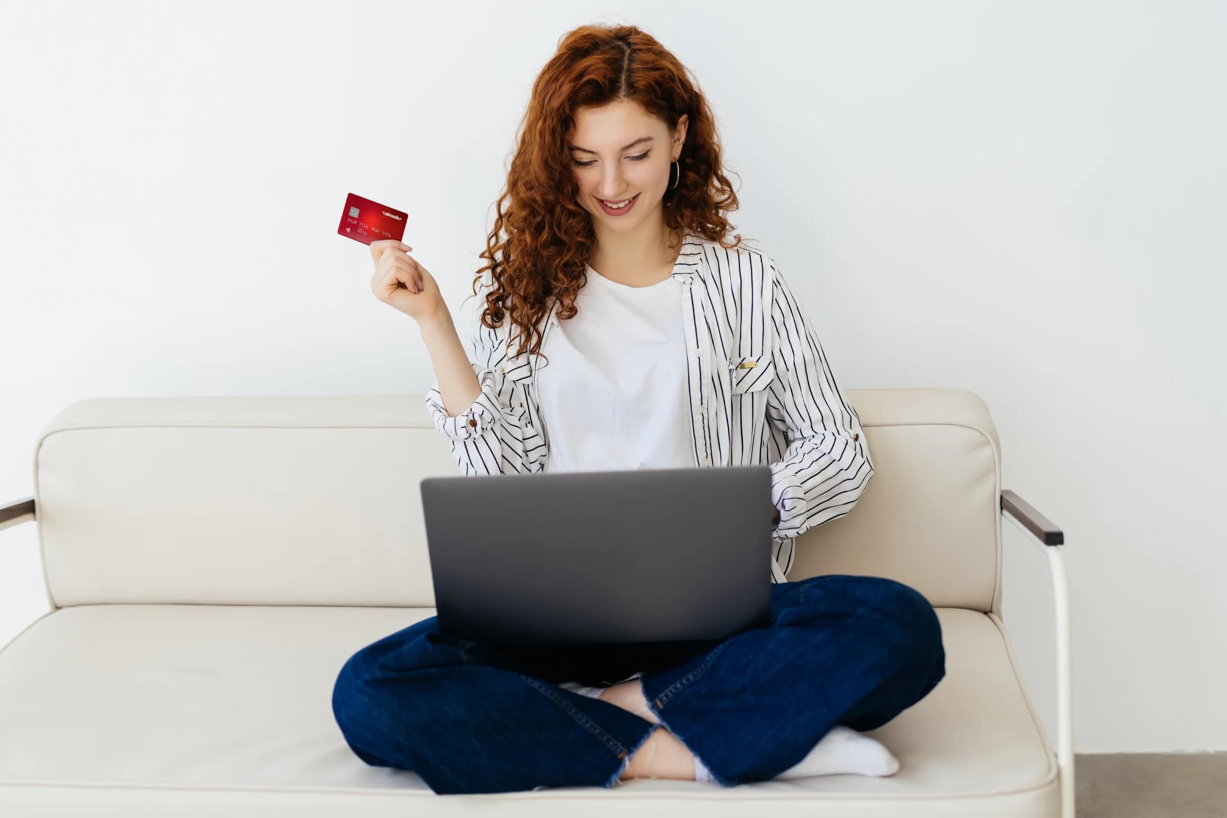 Woman with curly hair sits on a sofa using a laptop, holding a red credit card, wearing a striped shirt and jeans, smiling.