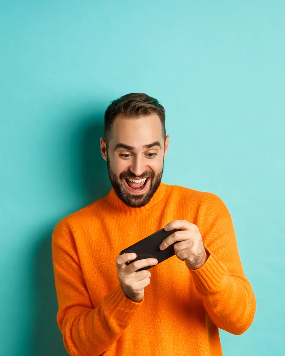 A man in an orange sweater looks excitedly at his smartphone against a turquoise background.