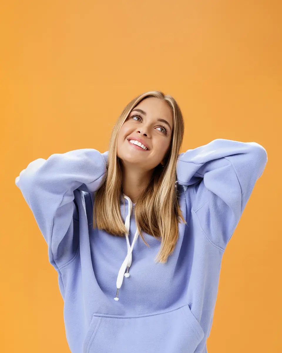 A woman in a light blue hoodie smiles and looks upward against an orange background, with her hands resting behind her neck.