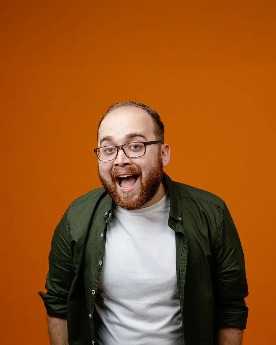 A bearded man wearing glasses, a white shirt, and a green jacket smiles widely against an orange background.
